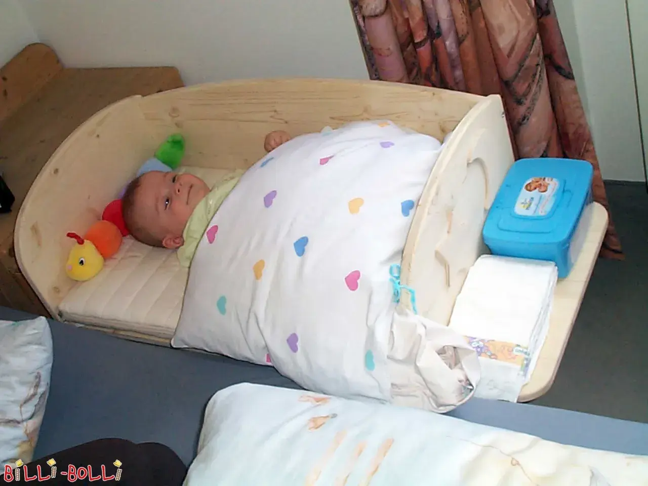 Ein süßes Baby schläft in einem selbstgebauten Babybett aus Holz. Neben ihm liegt ein bunter Raupen-Greifling. Windeln und Feuchttücher sind griffbereit.