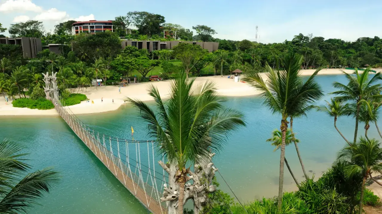Hängende Brücke über türkisfarbenem Wasser führt zu einem sonnigen singapur strand mit Palmen und Resorts im Hintergrund.