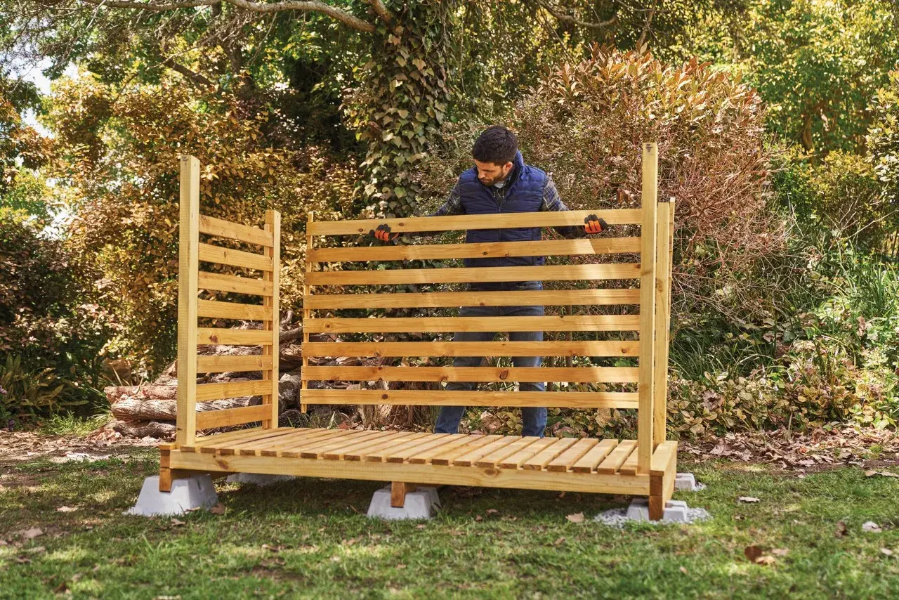 Uomo in piedi davanti a una nuova legnaia in legno, pronta per accogliere la legna per l'inverno.