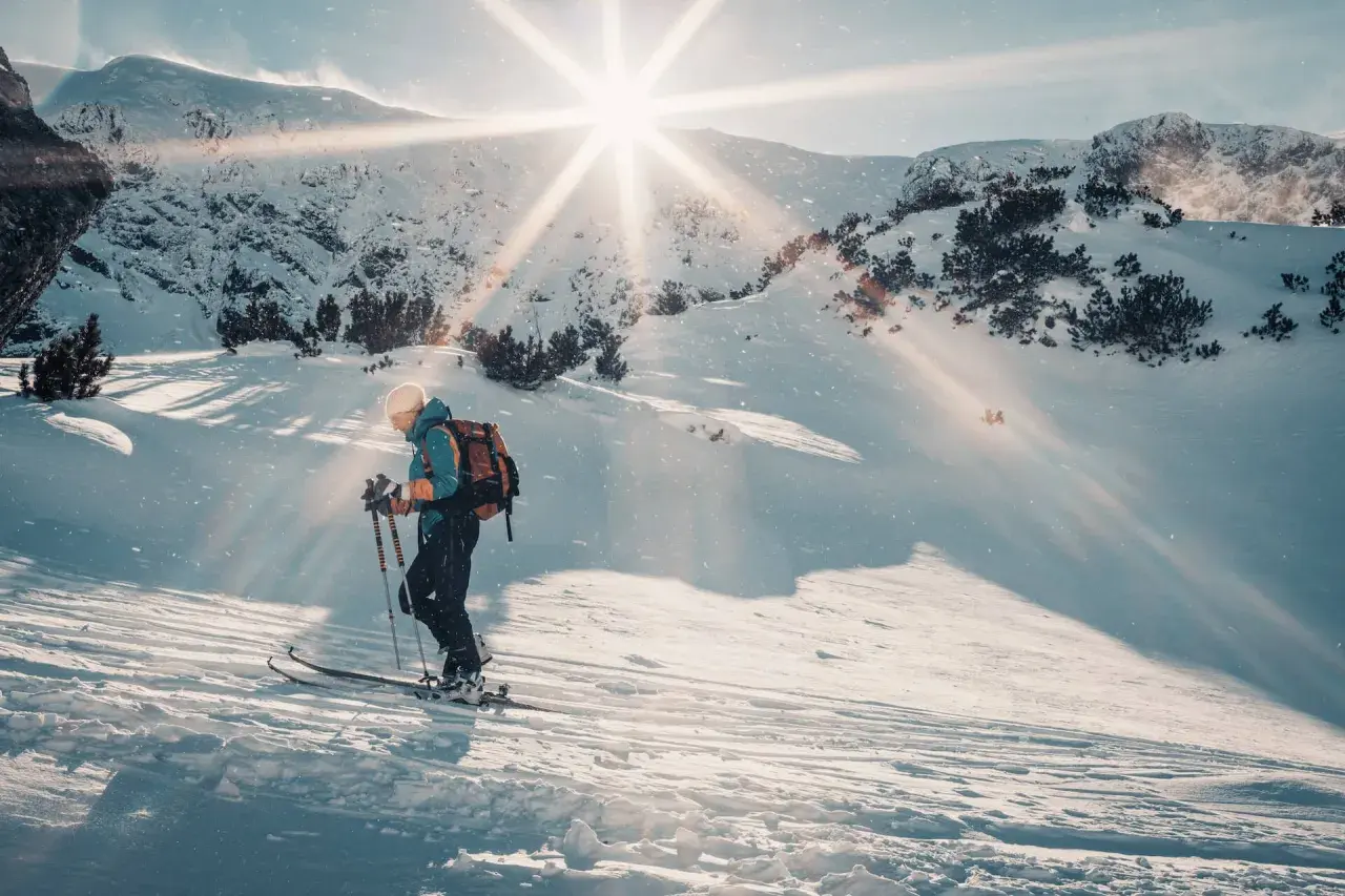 Skitouring co to? Narciarz z plecakiem wspina się po ośnieżonym stoku, słońce przebija się przez chmury, tworząc magiczną atmosferę.