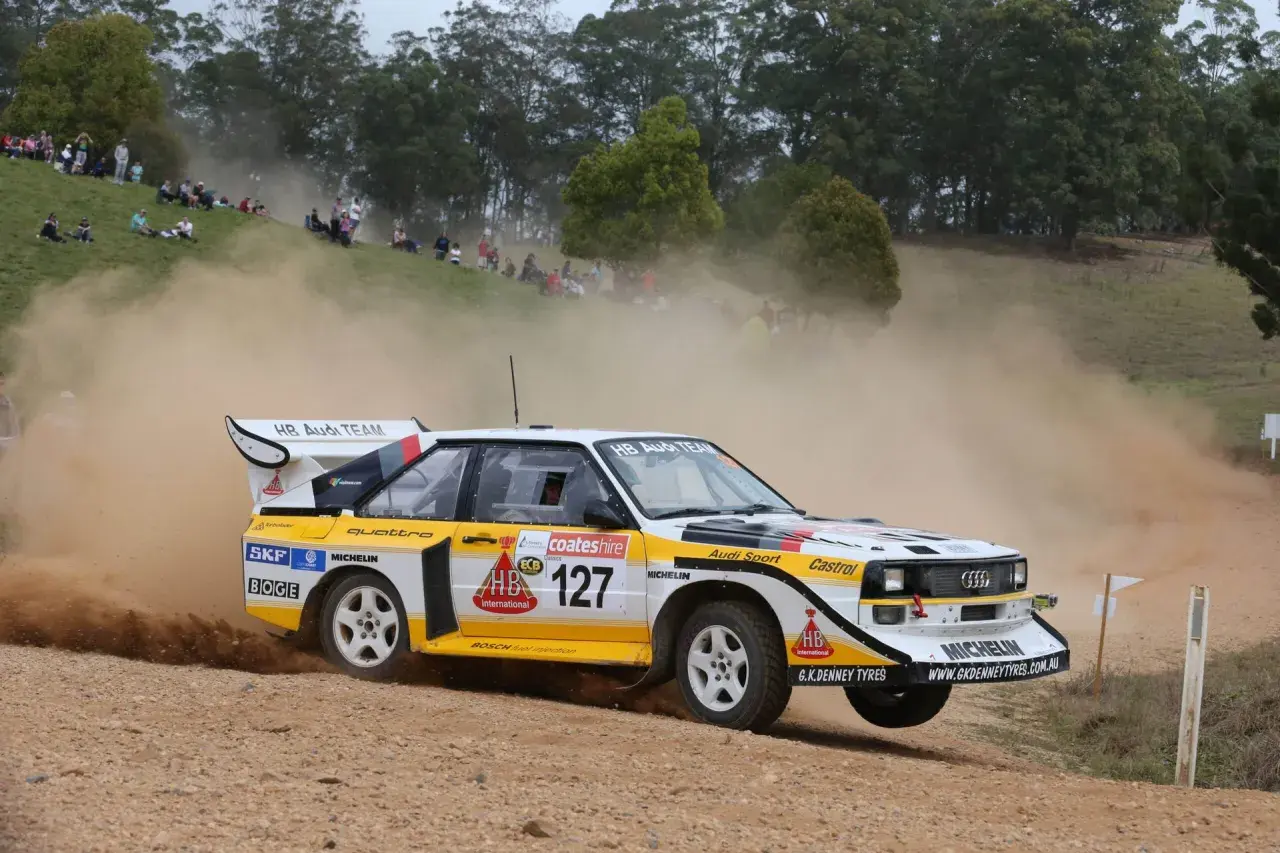 Klasyczny Audi Quattro z grupy B, oznaczony numerem 127, pędzi po szutrowej drodze, wzbijając tumany kurzu.