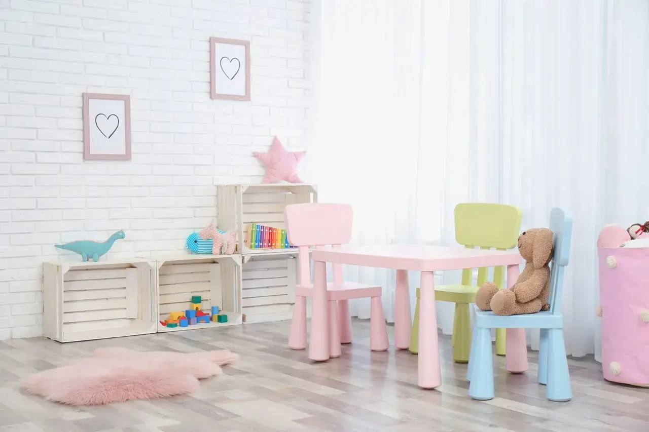 Kolorowy stolik z krzesełkami dla dzieci, idealny do zabawy i nauki. Na ścianie wiszą ramki z serduszkami, a obok stoją drewniane skrzynki z zabawkami.