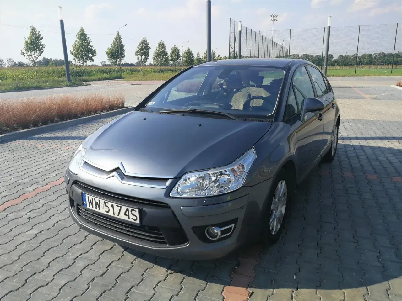 Szary Citroen C4 z 2008 roku na parkingu z kostki brukowej, w tle drzewa i boisko.
