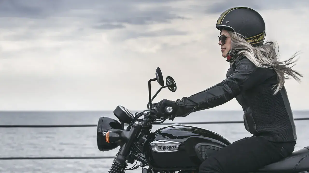 Mujer rubia en moto Triumph negra, con casco y chaqueta de cuero, disfrutando del paisaje. Las motos Triumph se fabrican con pasión.