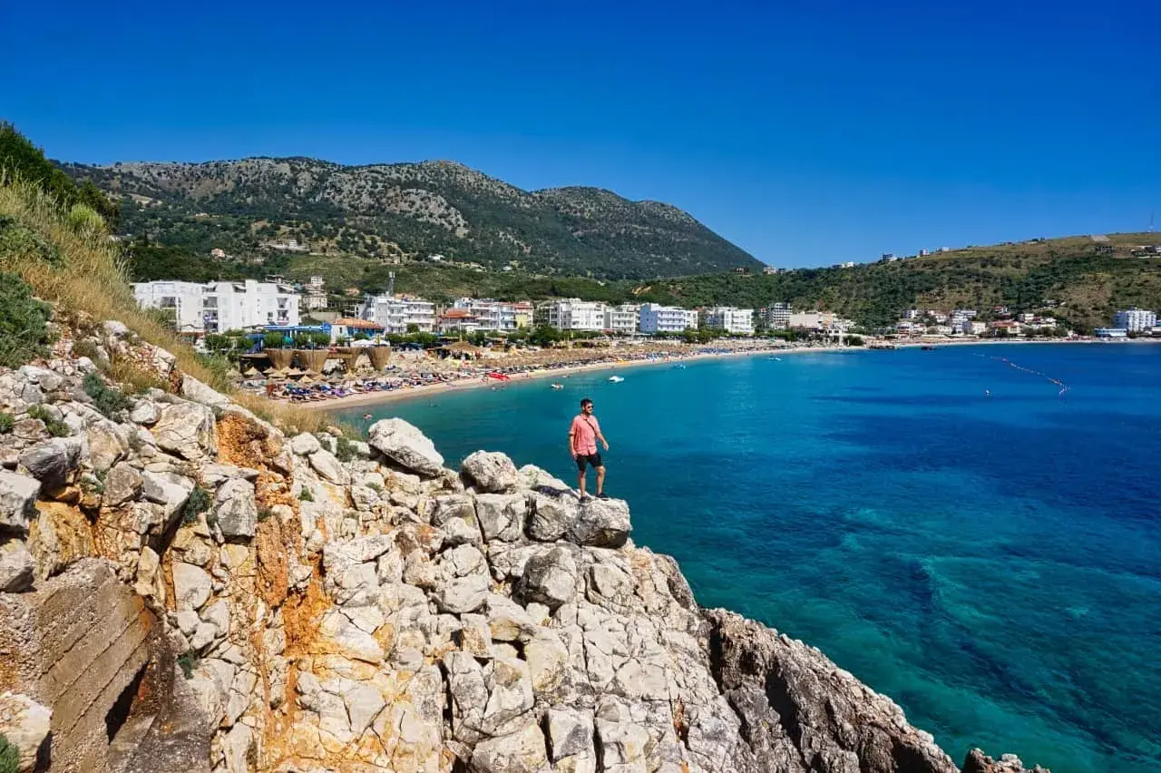 Mann steht auf Felsen mit Blick auf eine Bucht mit Strand und Hotels. Perfekt, um zu wissen, wo in Albanien Urlaub machen!