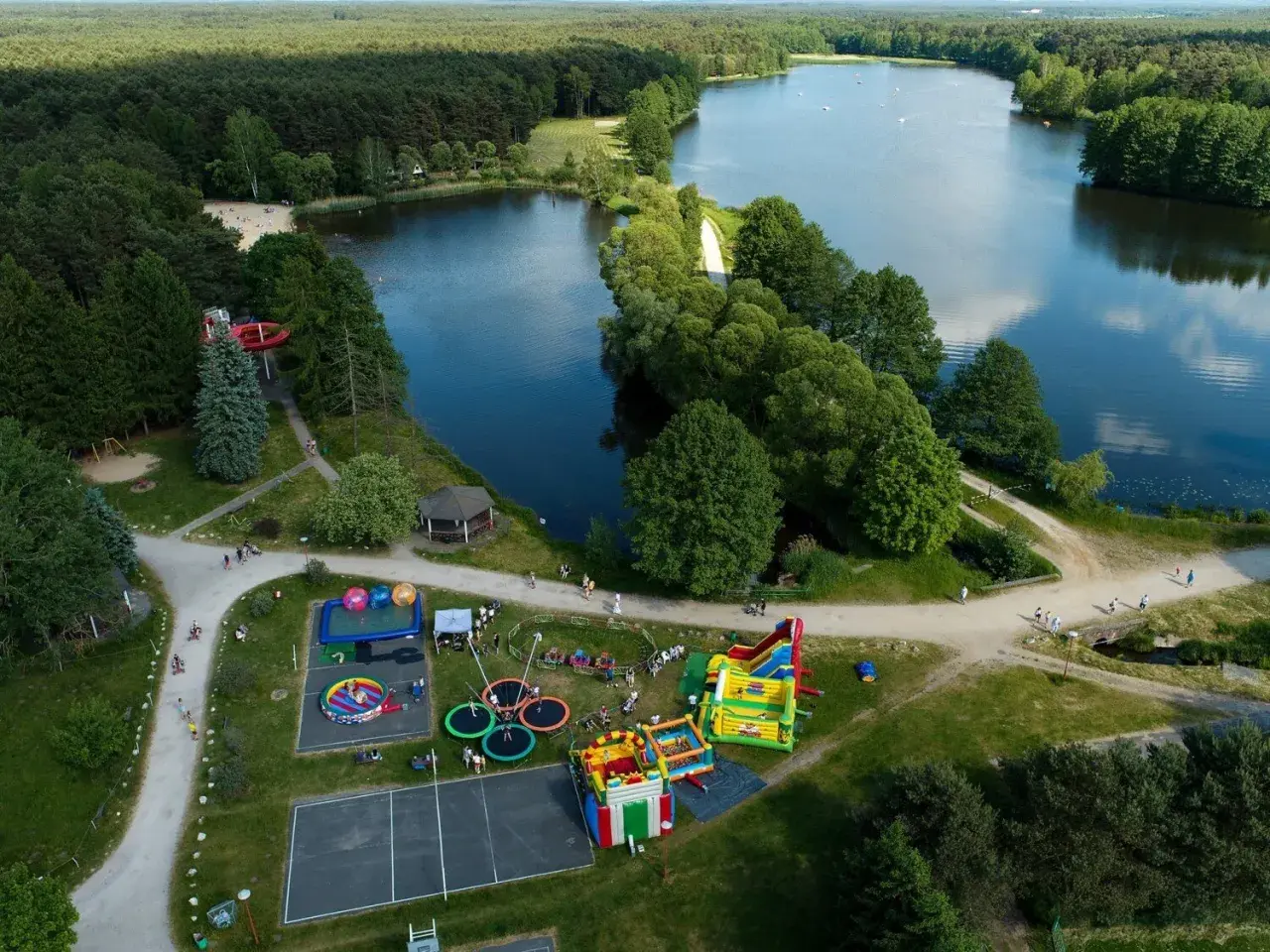 Rekreacja nad jeziorem w Bełchatowie: plac zabaw z dmuchańcami, trampolinami i zjeżdżalnią wodną, otoczony lasem i wodą.