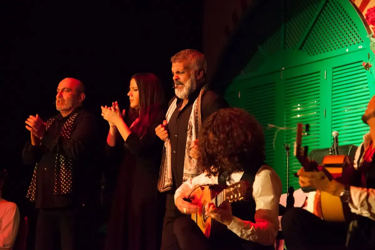 Artistas flamencos, incluyendo cantaores flamencos famosos, aplauden y tocan la guitarra en un escenario con una puerta verde de fondo.