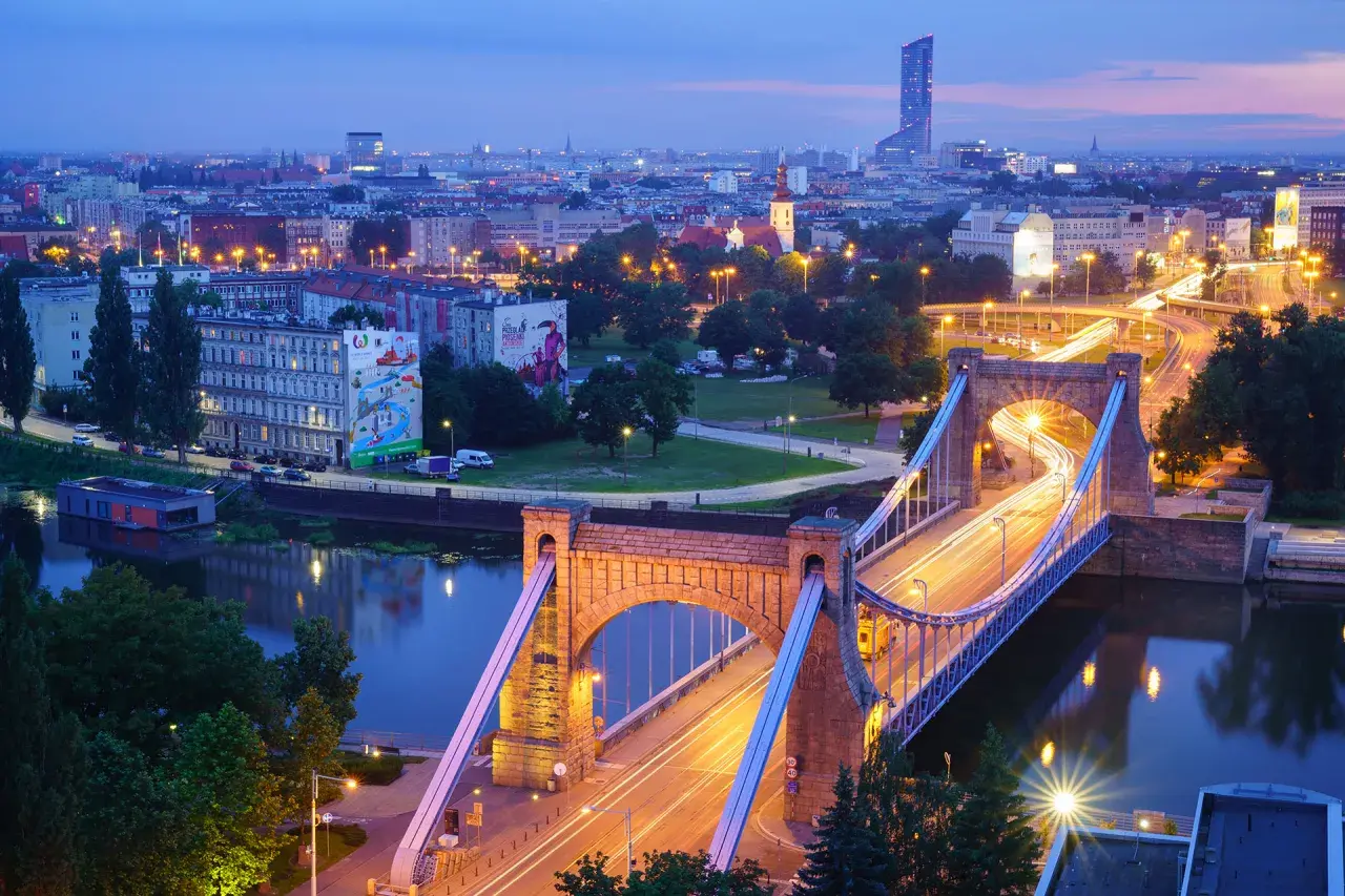 Most Wrocławia w niebieskiej godzinie w fotografii. Most Grunwaldzki i miejska panorama nocą, z rozmazanymi światłami samochodów tworzącymi smugi.