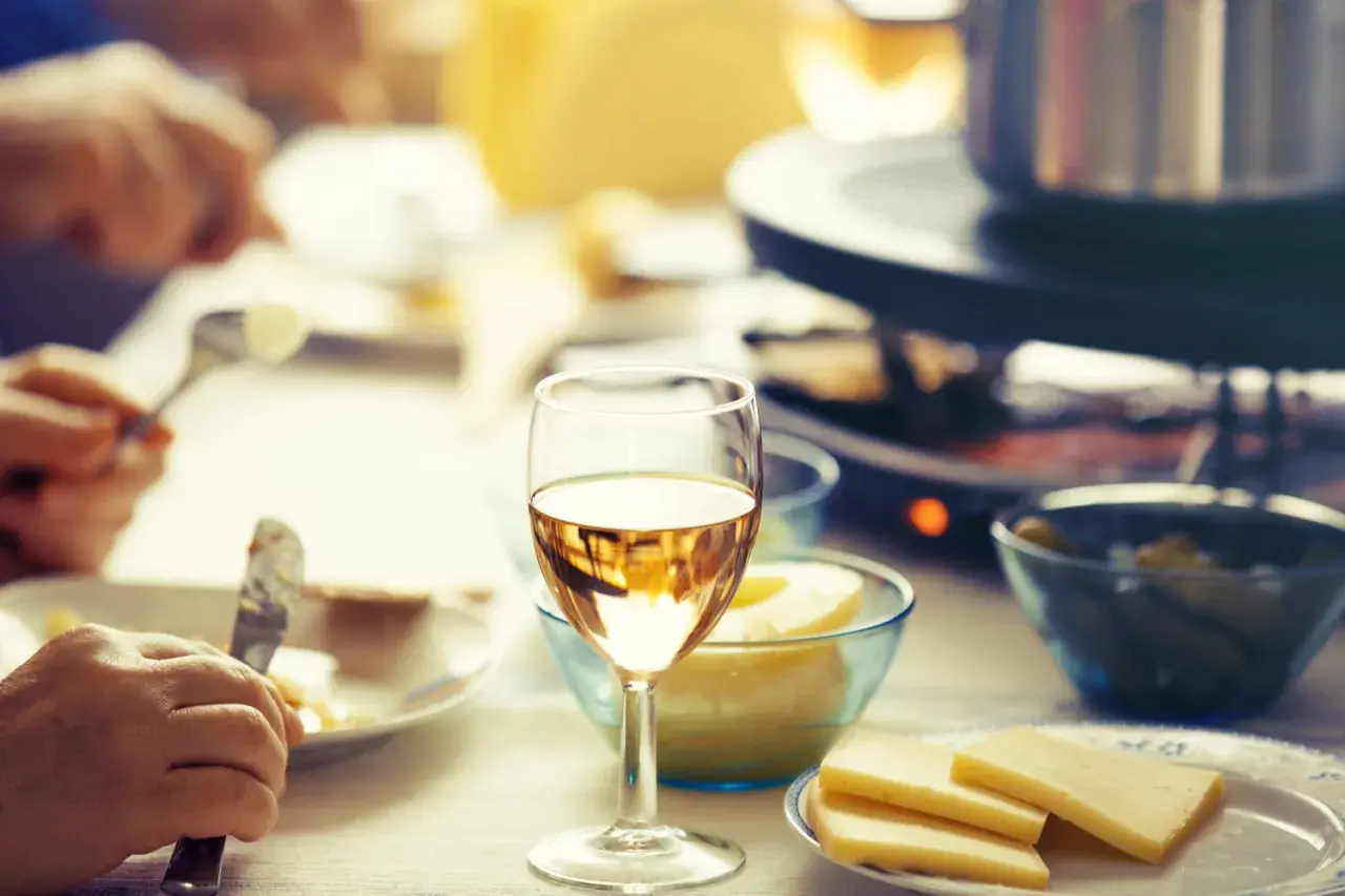 Un verre de vin blanc sec ou moelleux accompagne une raclette conviviale. Des morceaux de fromage et des cornichons sont prêts à être dégustés.