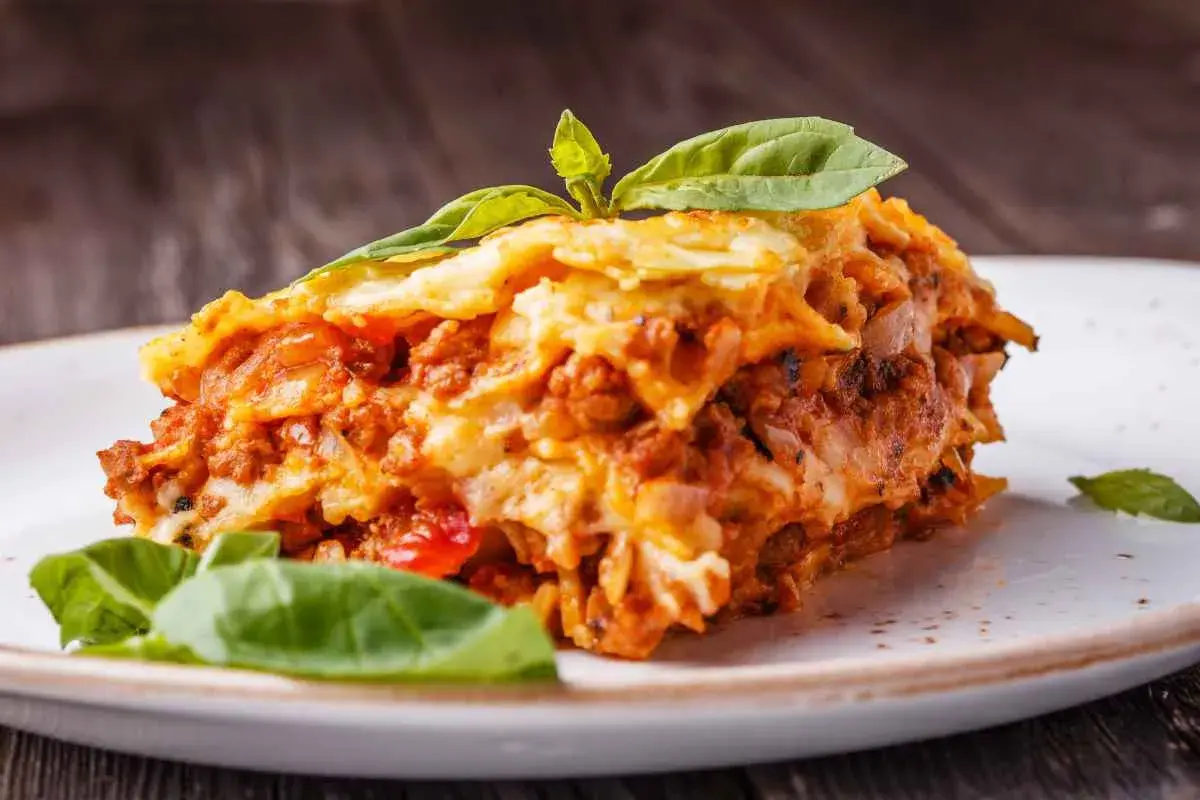 Una fetta di lasagna salentina, ricca e saporita, guarnita con basilico fresco, su un piatto bianco.