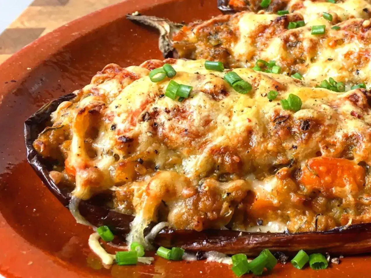 Berenjenas rellenas gratinadas con queso derretido y cebolleta picada, servidas en una cazuela de barro.