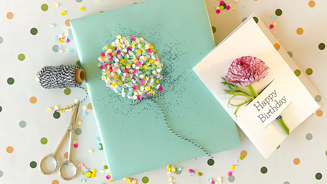 Ein liebevoll gestaltetes Geschenk zum 1. Geburtstag: Geschenkpapier mit Glitzer-Ballon, Schnur, Schere und eine Karte mit Blüte.