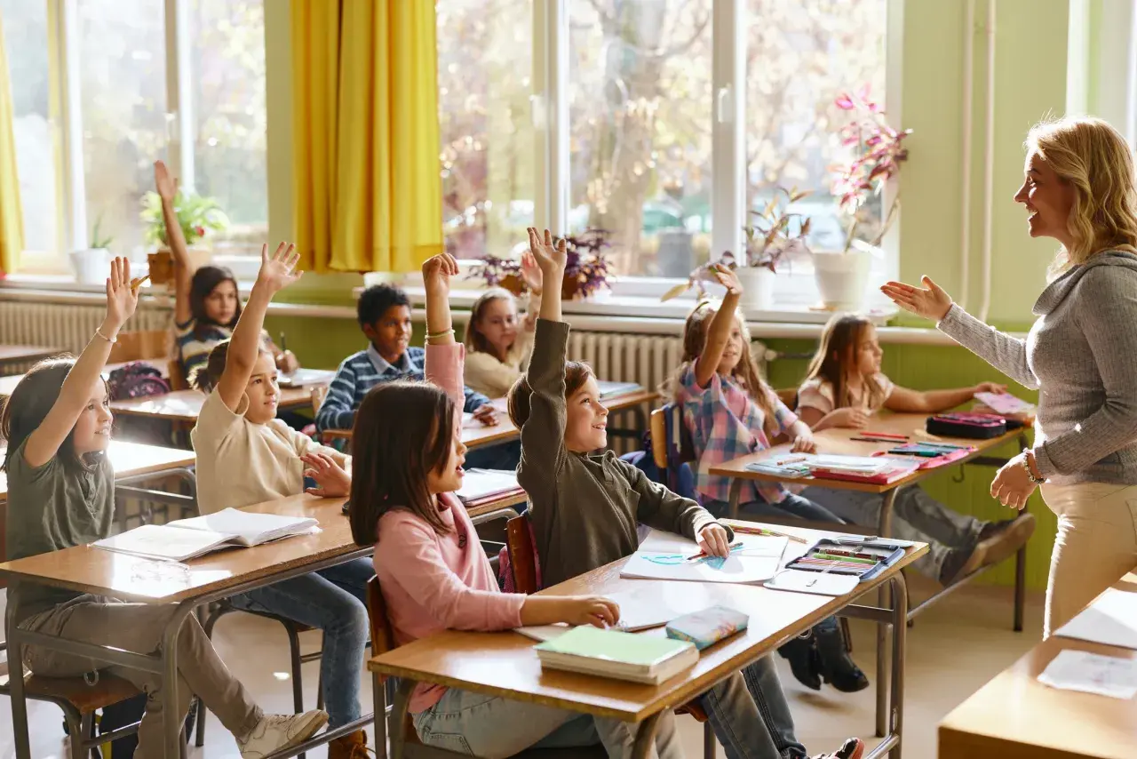 Grundschüler mit erhobenen Händen, bereit zu antworten. Die Sitzordnung zur Wand hin ist gut erkennbar, die Lehrerin lächelt.