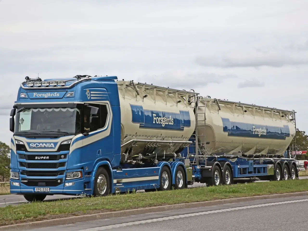 Ein blauer Scania-Sattelzug mit zwei Siloanhängern, beladen für Forsgårds, fährt auf einer schwedischen Straße. Das Tempolimit in Schweden ist zu beachten.