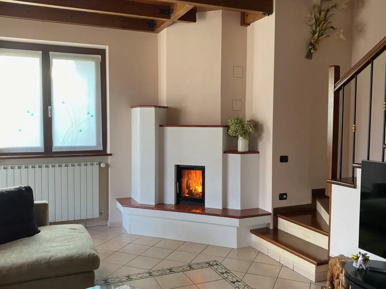 Camino moderno con fiamme vive. La struttura in **calce resiste alle alte temperature**, creando un angolo accogliente con scala e divano.