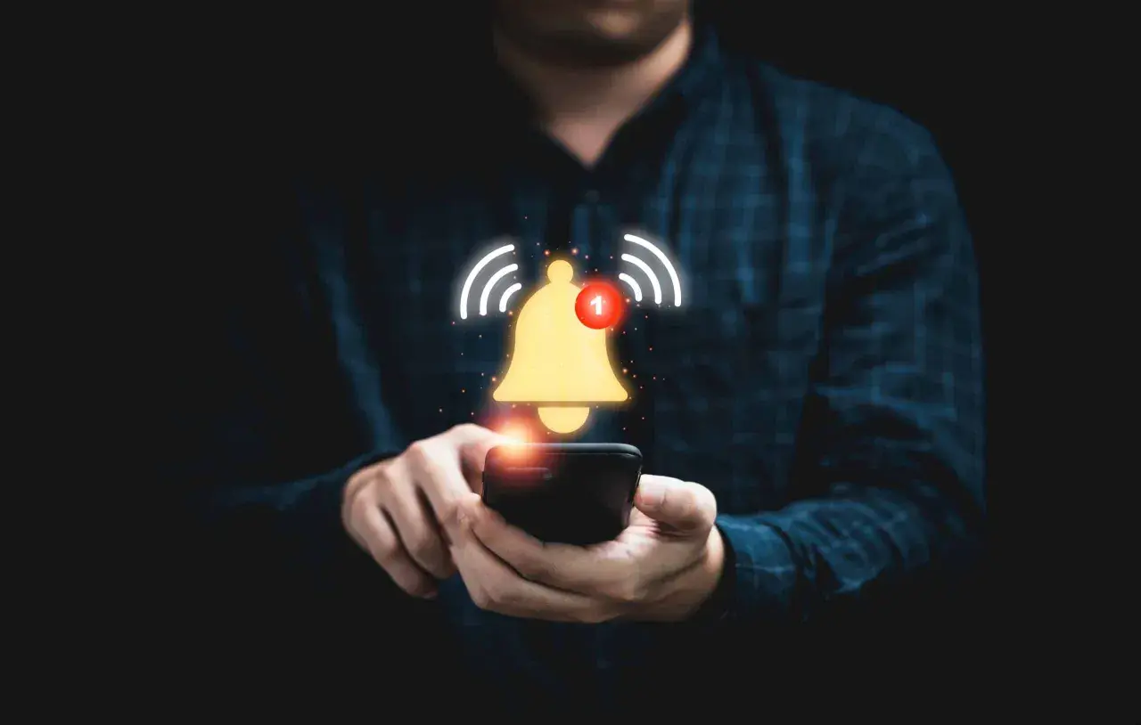 Dłoń trzymająca smartfon, nad którym unosi się złoty dzwonek z powiadomieniem.