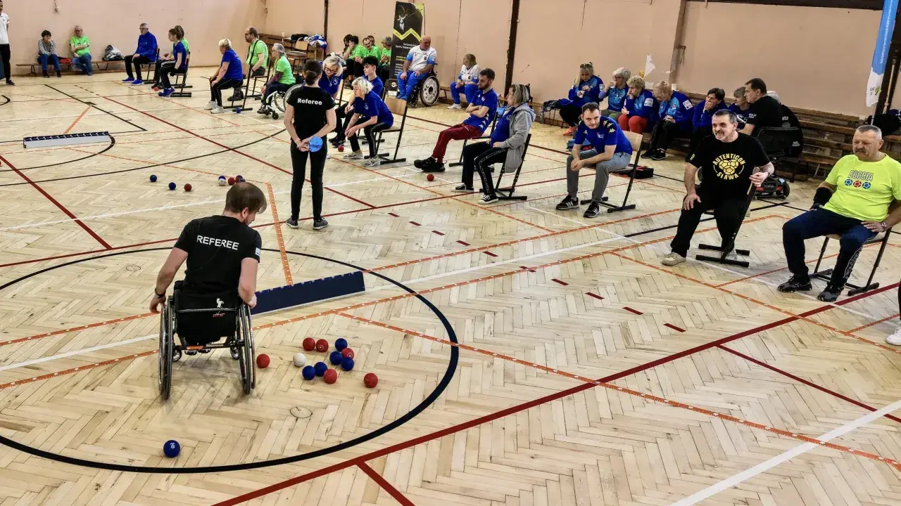 Zawodnicy grają w boccię, sportową grę precyzyjną. Na boisku widać kolorowe kule i zawodnika na wózku inwalidzkim.