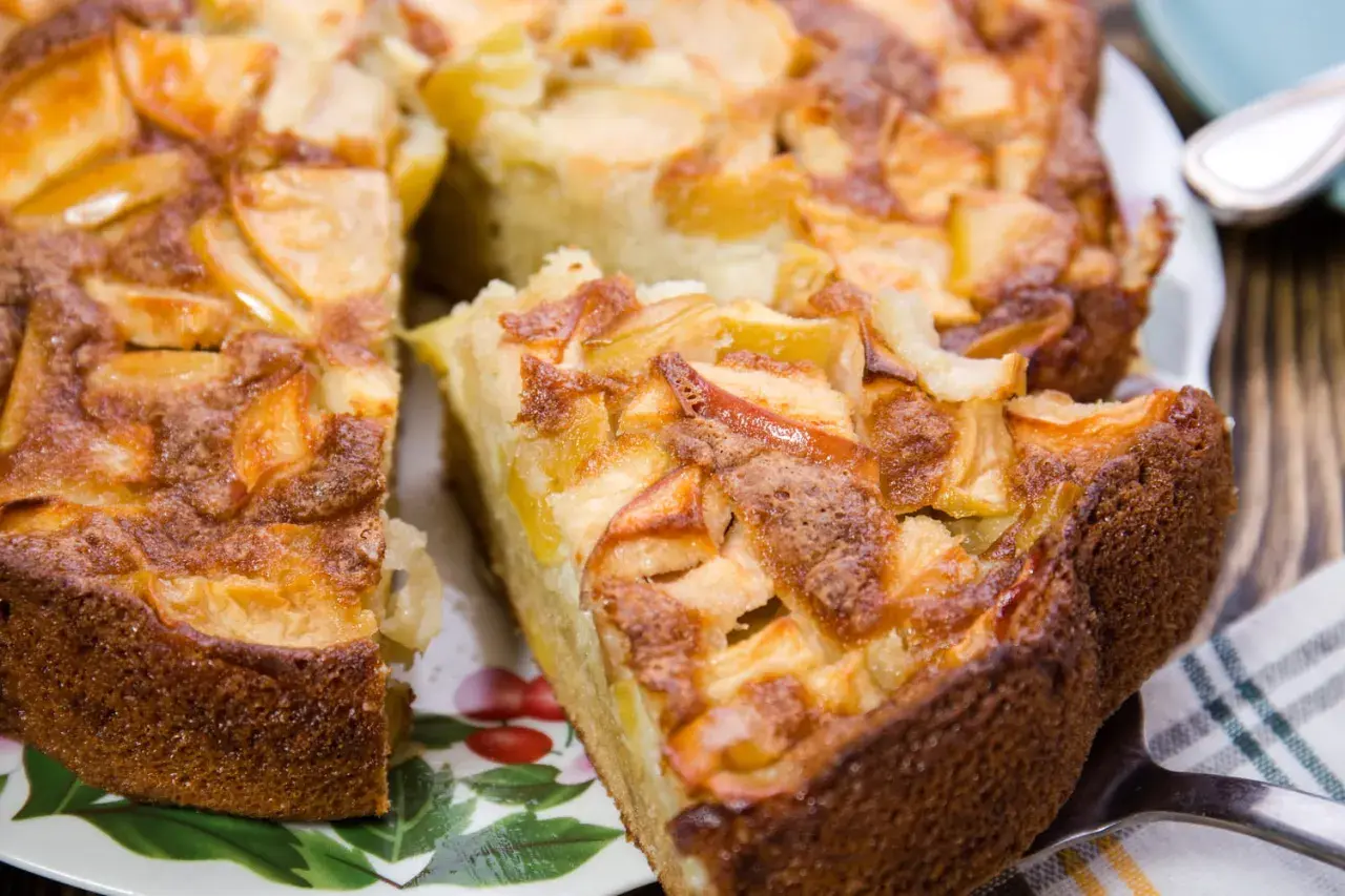 Un délicieux gâteau aux pommes, doré et moelleux, avec des morceaux de fruits caramélisés sur le dessus. Une part est déjà coupée, prête à être dégustée.