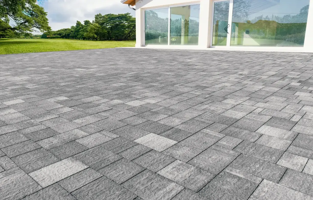 Un ampio patio in gres carrabile grigio, con pavimentazione a motivi geometrici, si estende davanti a una moderna casa con vetrate.