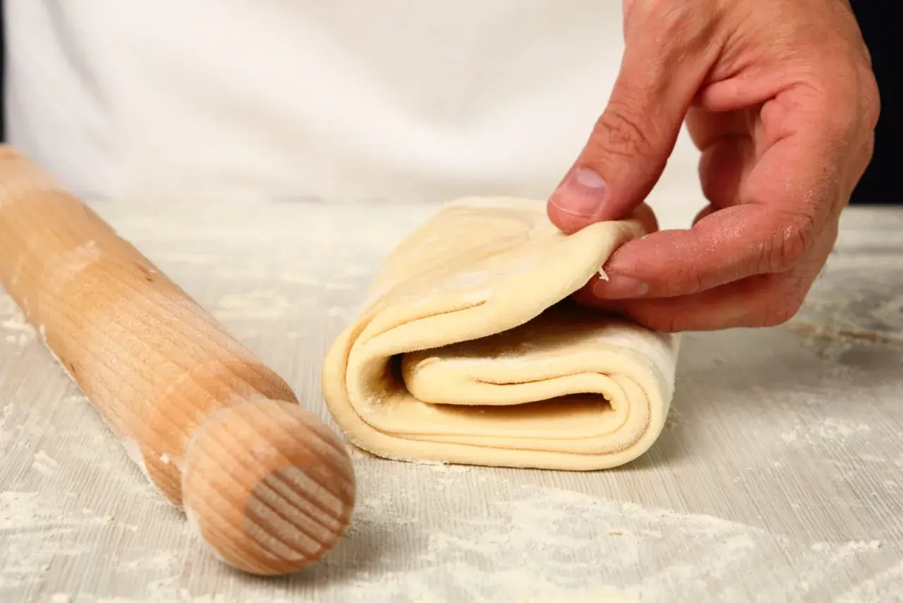 Mani che piegano pasta sfoglia, pronta per diventare pasta fatta in casa. Un mattarello è accanto, pronto per il prossimo passaggio.