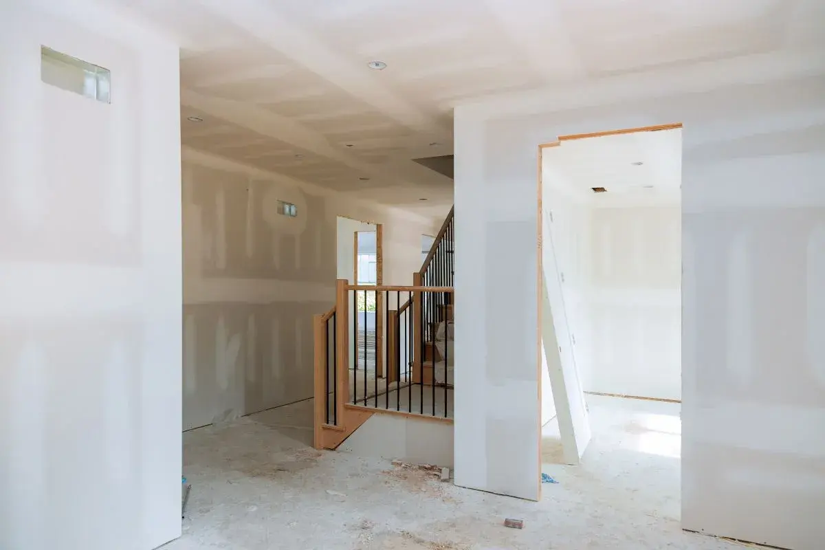 Interno di una casa in costruzione con pareti in cartongesso grezzo, una scala in legno e metallo e un'apertura verso un'altra stanza.