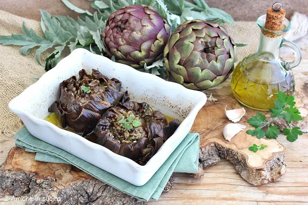Carciofi al forno morbidi ripieni, serviti in una teglia bianca con olio d'oliva, aglio e prezzemolo.