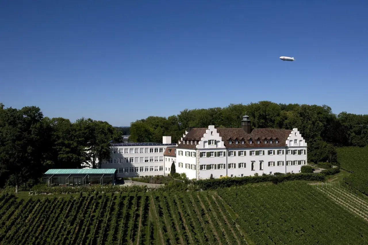 Schloss Hersberg, umgeben von Weinbergen, mit einem Zeppelin am blauen Himmel.