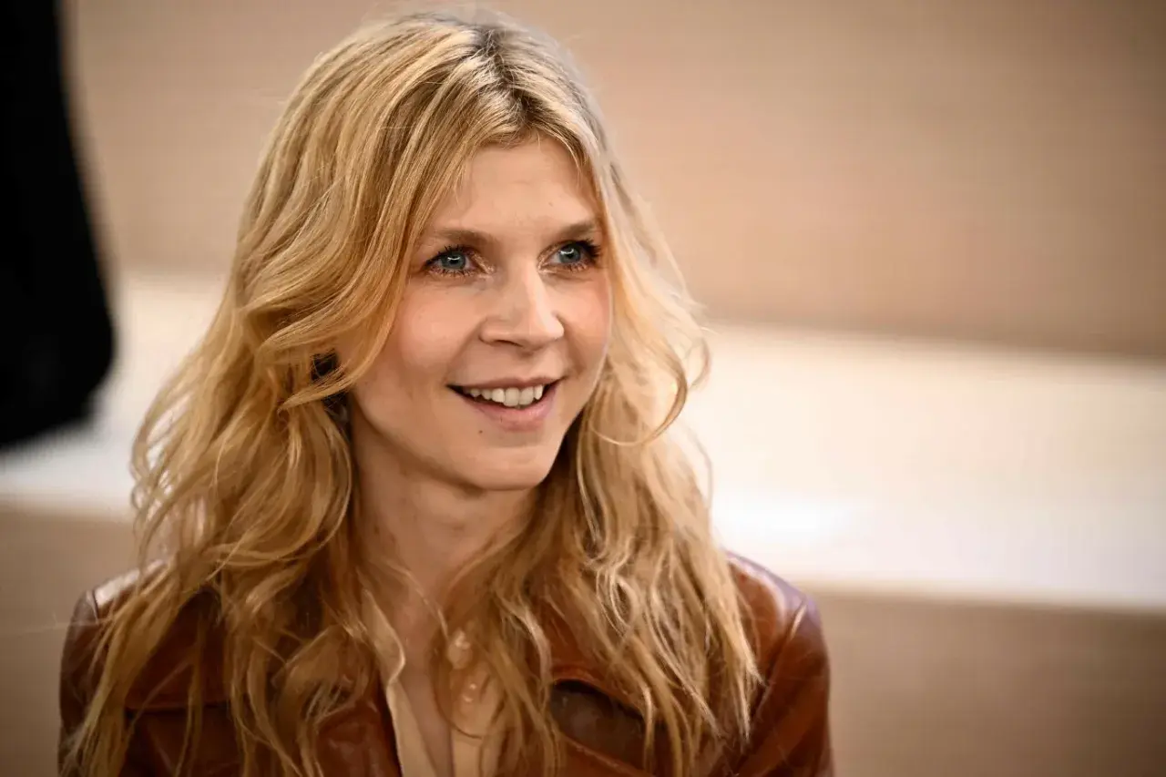 Uśmiechnięta Clémence Poésy z falującymi blond włosami i niebieskimi oczami, ubrana w brązową skórzaną kurtkę.