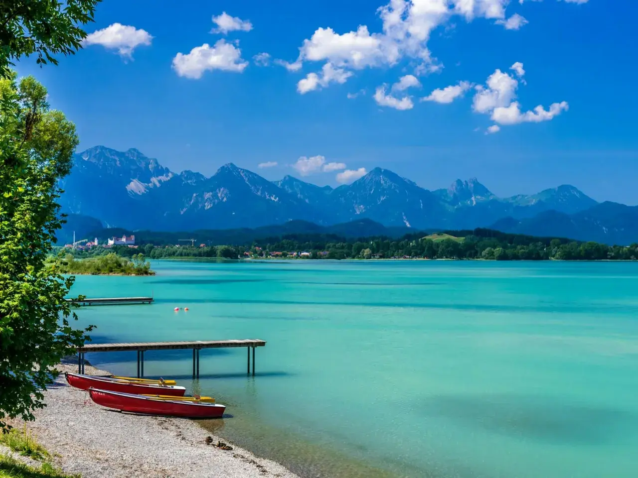 Türkisblaues Wasser am Badesee Bayern, mit roten Ruderbooten am Kiesstrand und majestätischen Alpen im Hintergrund.