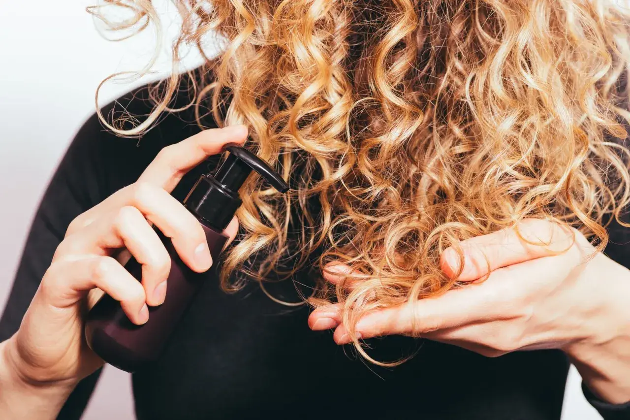Delikatna pielęgnacja włosów wysokoporowatych. Ręka z buteleczką kosmetyku aplikuje go na blond loki.