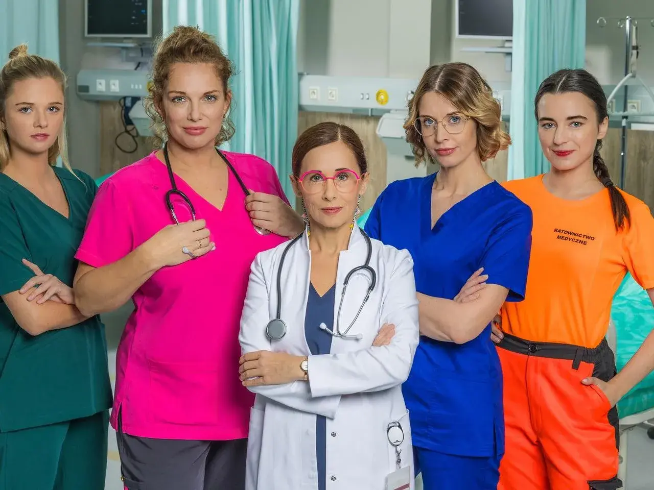 Pięć kobiet w strojach medycznych, obsada serialu "Szpital św. Anny", pozuje do zdjęcia w szpitalnej sali.