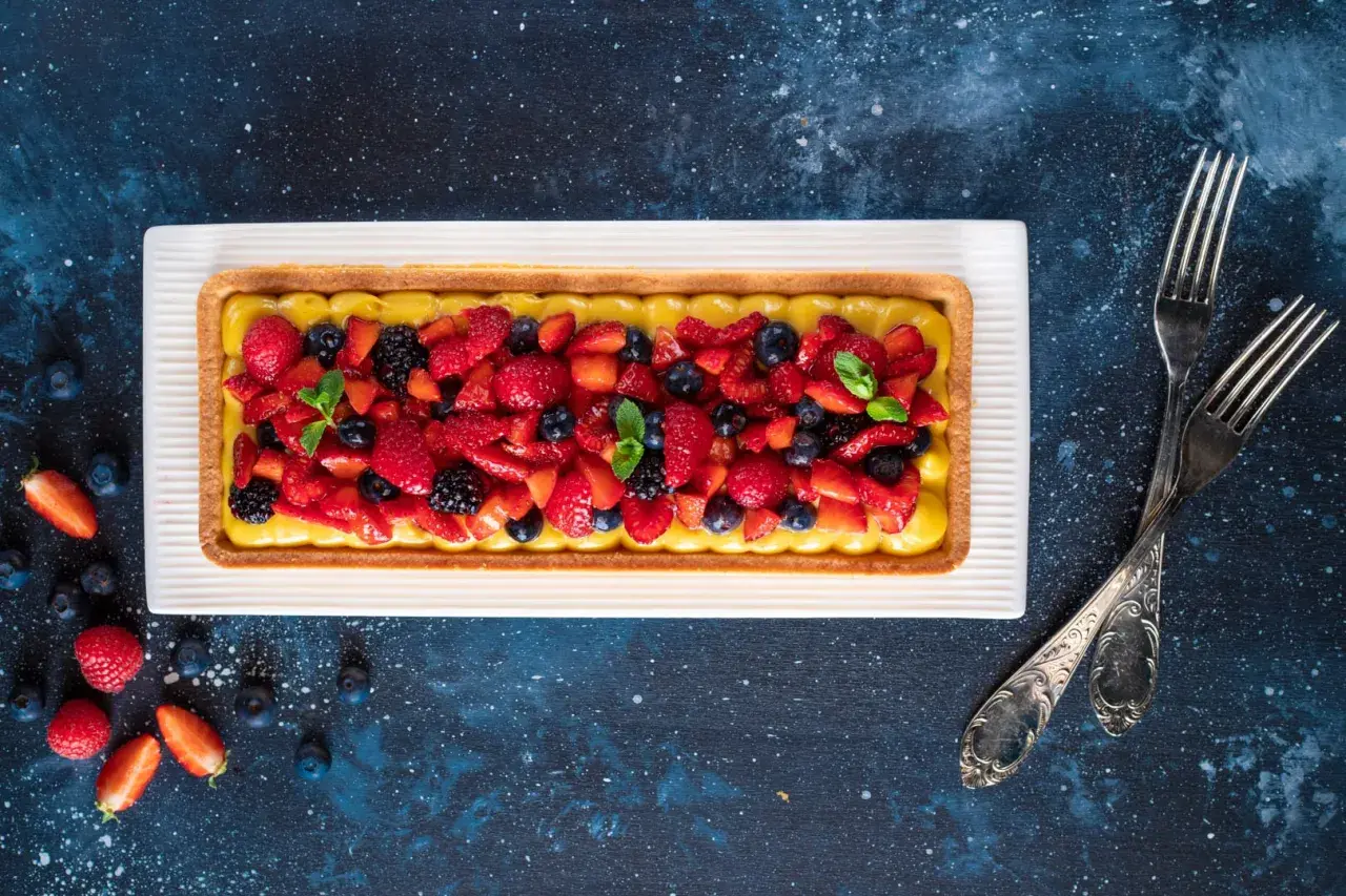Una crostata di frutta moderna, con crema gialla e un tripudio di fragole, mirtilli e more fresche, guarnita con menta.