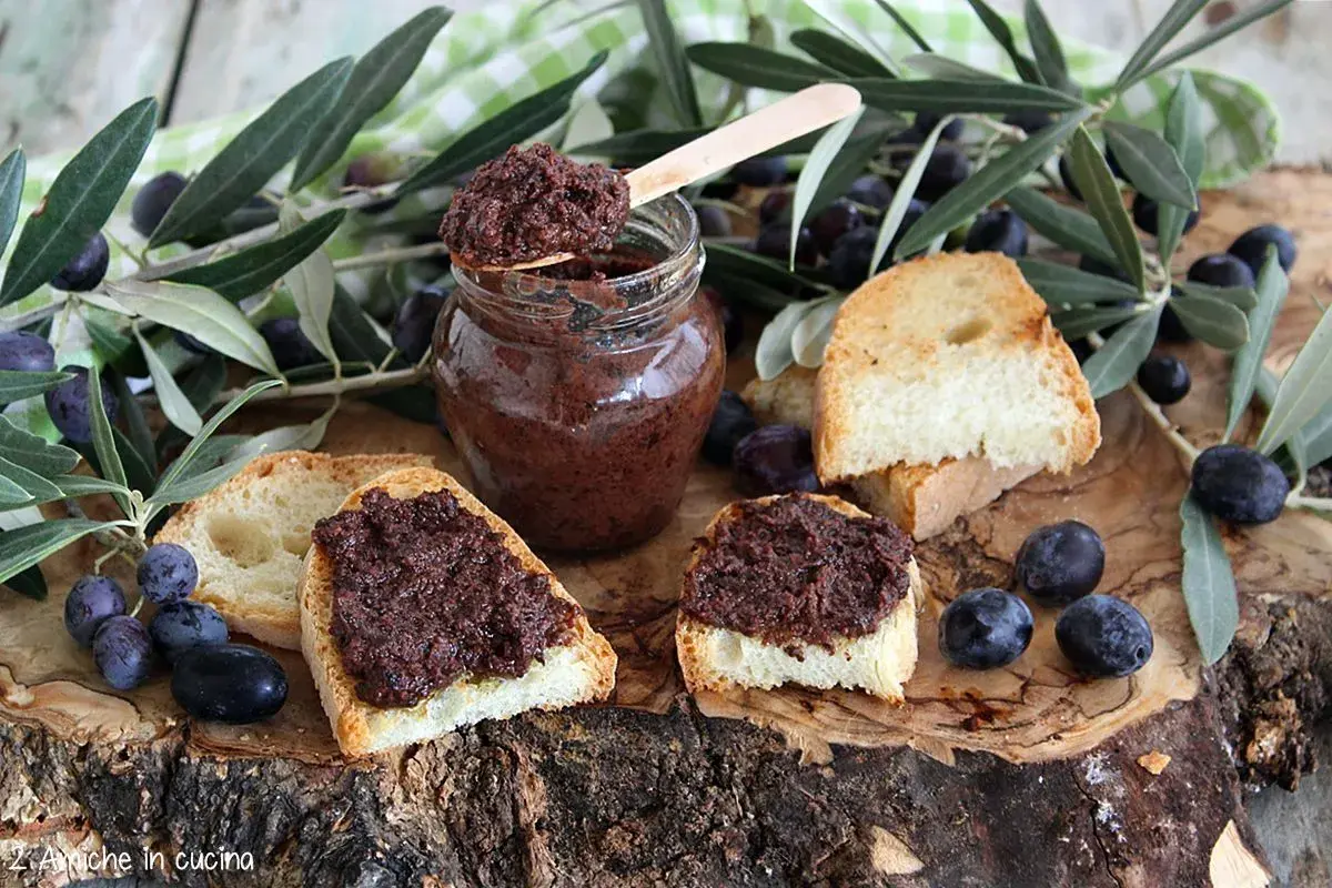 Ecco come si fa il patè di olive: bruschette con paté scuro, olive nere e rametti d'ulivo su un tagliere di legno.