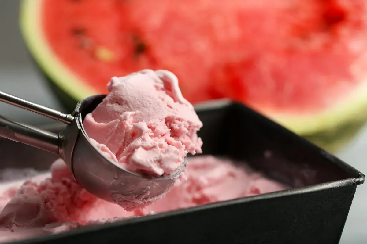 Un cucchiaio raccoglie una pallina di cremoso gelato anguria fatto in casa, con una fetta di anguria sullo sfondo.