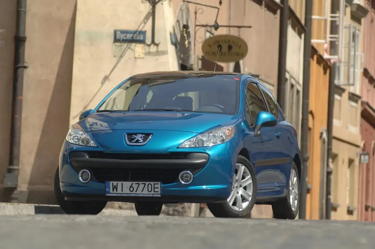 Błękitny Peugeot 207 z wyraźnymi żarówkami reflektorów stoi na brukowanej ulicy.