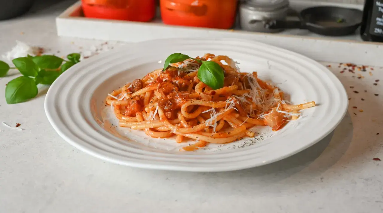En tallrik med bucatini pasta i tomatsås, toppad med riven parmesan och färsk basilika. Ett perfekt bucatini pasta recept för en god middag.