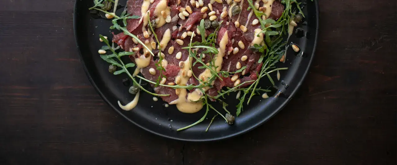 Rinder Carpaccio mit Pinienkernen, Kapern und Rucola auf schwarzer Platte, beträufelt mit einer cremigen Sauce.