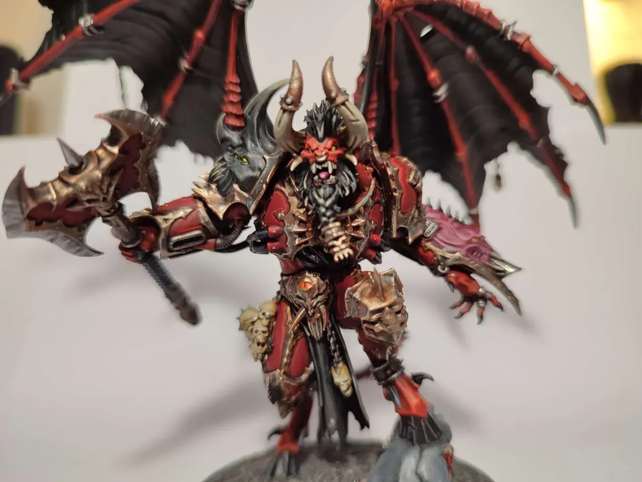 Groźny chaos daemon prince z czerwono-złotą zbroją, czarnymi skrzydłami i toporem.