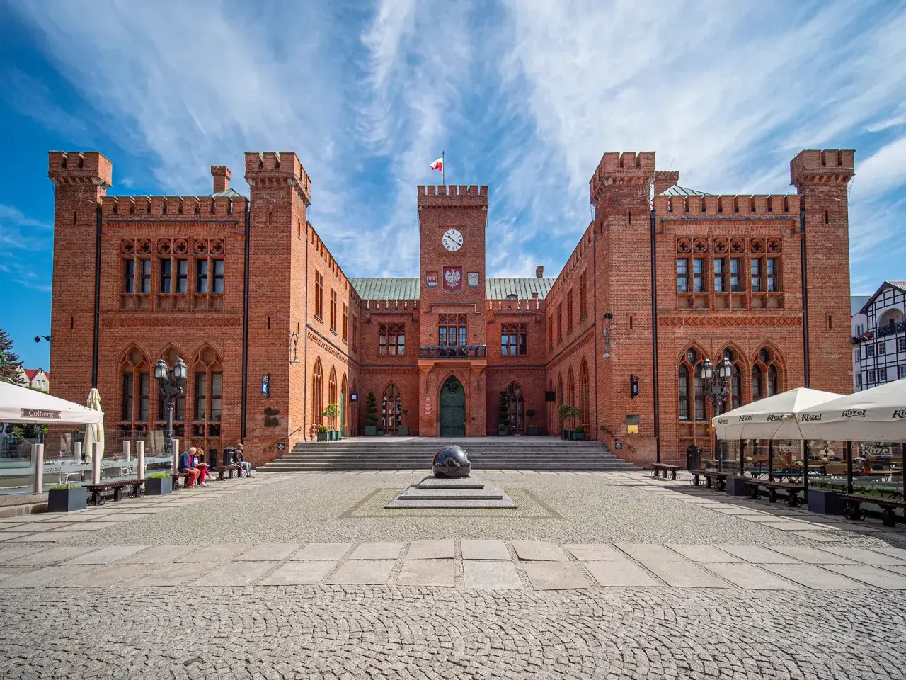 Rynek Kołobrzeg: ceglany ratusz z zegarem i flagą, przed nim kamienna rzeźba i brukowany plac.