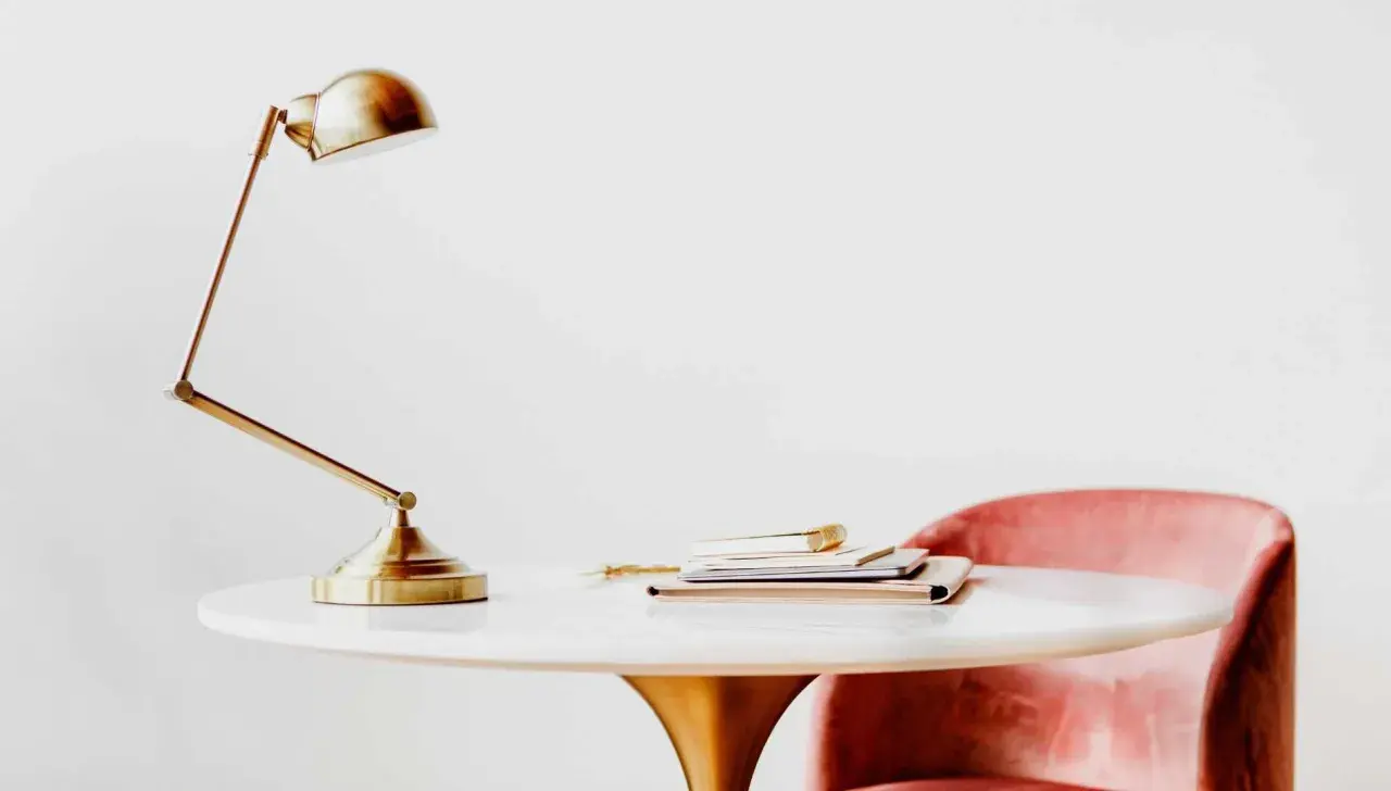 Elegante lampara de mesa metal dorada sobre un escritorio blanco con libros y una silla rosa.