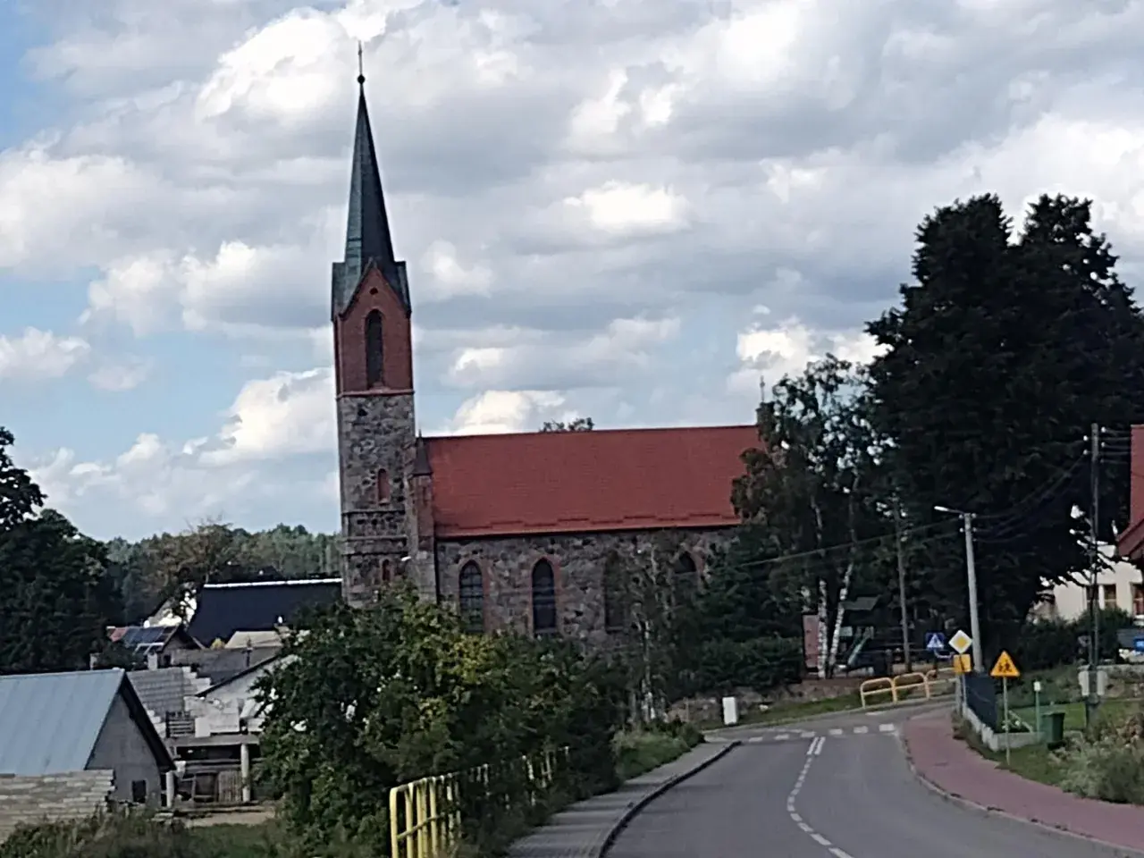 Kamienny kościół z czerwoną dachem i strzelistą wieżą to jedna z głównych atrakcji Sierakowic. Wokół widać domy i zieleń.