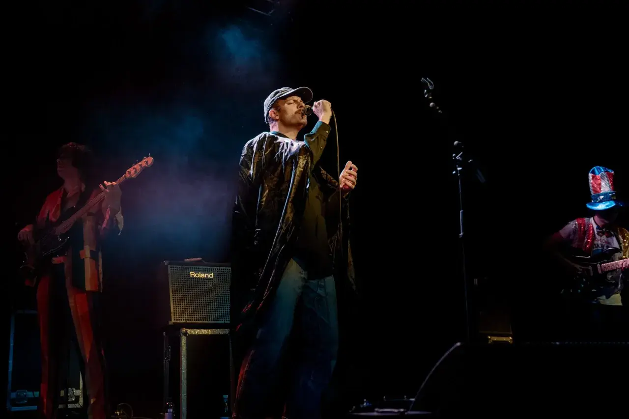 Mac DeMarco sjunger på scen i Stockholm, iklädd keps och en glansig jacka. En basist och en gitarrist syns också.