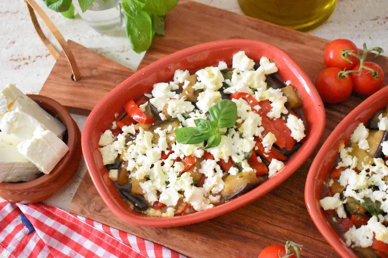 Pyszna pieczona feta z warzywami, podana w glinianym naczyniu, z dodatkiem świeżej bazylii i pomidorków.