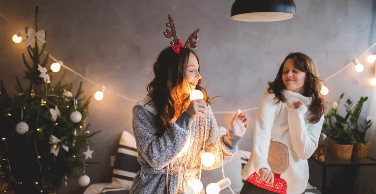 Due ragazze cantano canzoni di Natale in inglese con testo, tenendo una lampadina come microfono, circondate da luci natalizie e un albero addobbato.