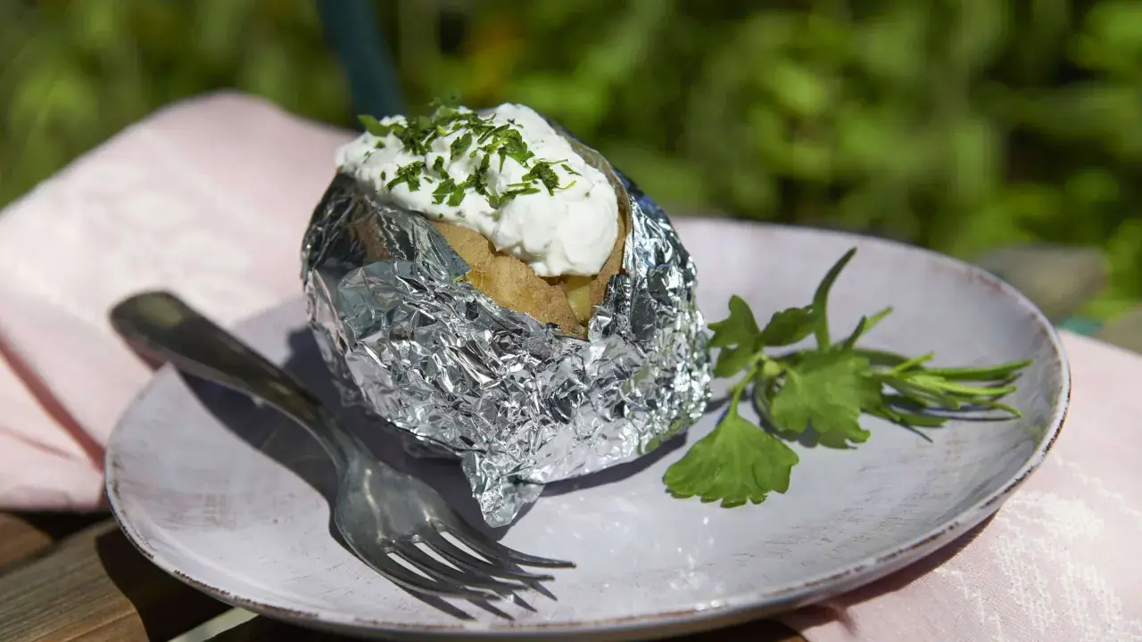 Folienkartoffeln vorkochen: Eine Kartoffel in Alufolie, belegt mit Quark und Kräutern, auf einem Teller mit Gabel und Petersilie.