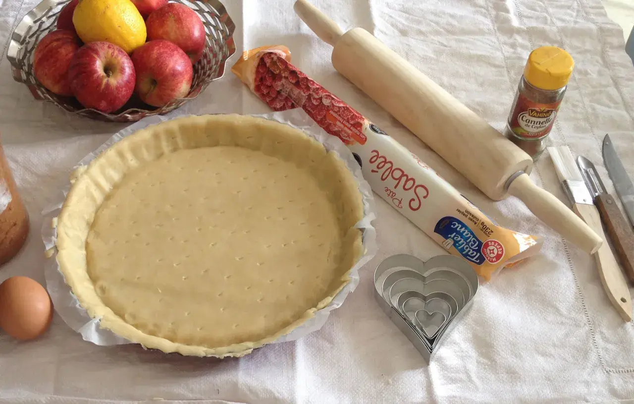 Préparation d'une tarte aux pommes simple : pâte sablée, pommes, citron, œuf, rouleau, emporte-pièces cœur et cannelle.