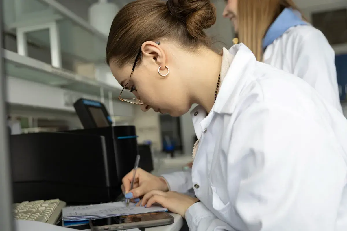 Studentka w białym fartuchu laboratoryjnym pisze notatki w zeszycie, obok klawiatury i telefonu.