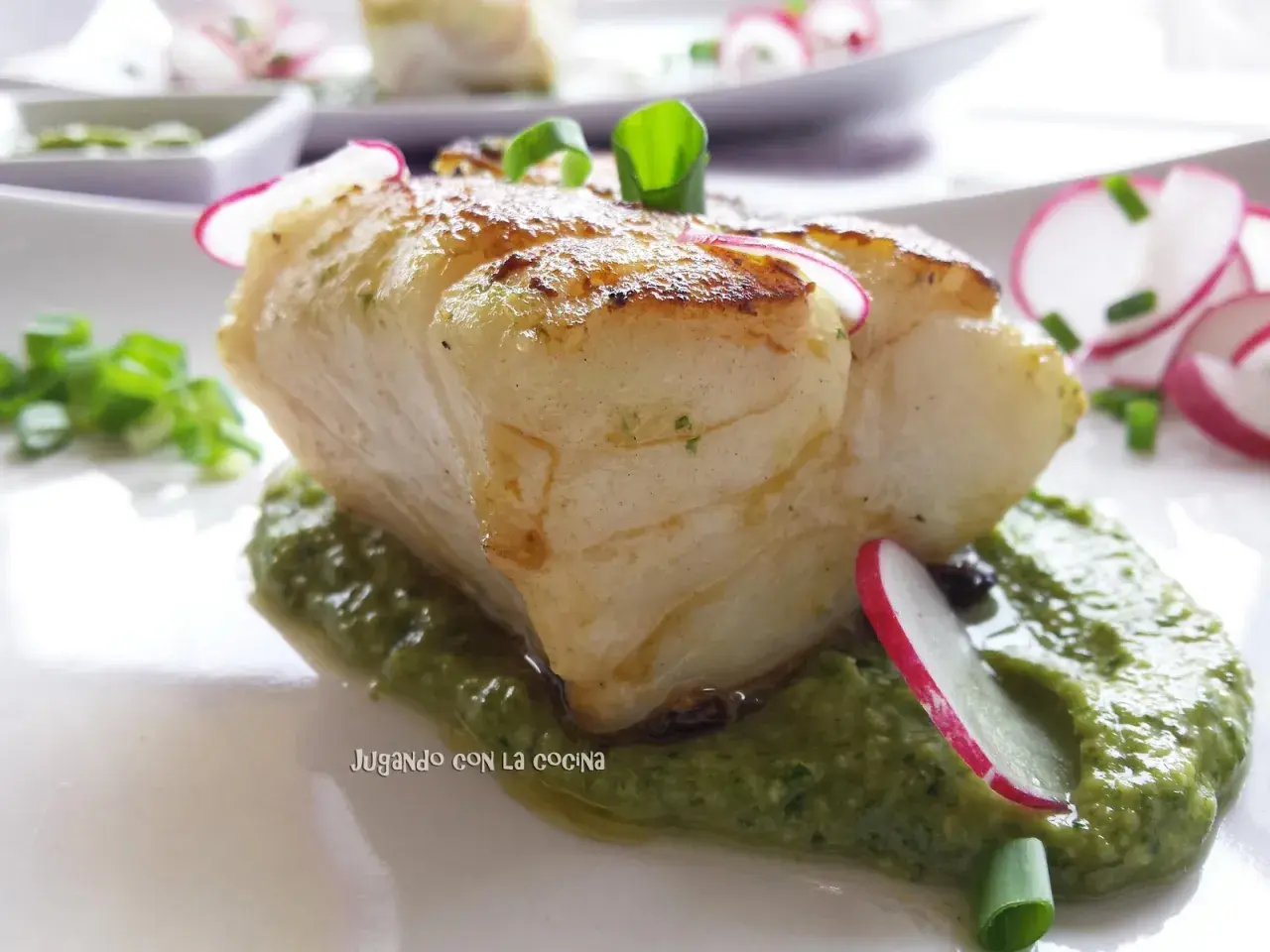 Un trozo de pescado blanco y tierno reposa sobre una cama de vibrante mojo verde, adornado con rodajas de rábano y cebolleta.