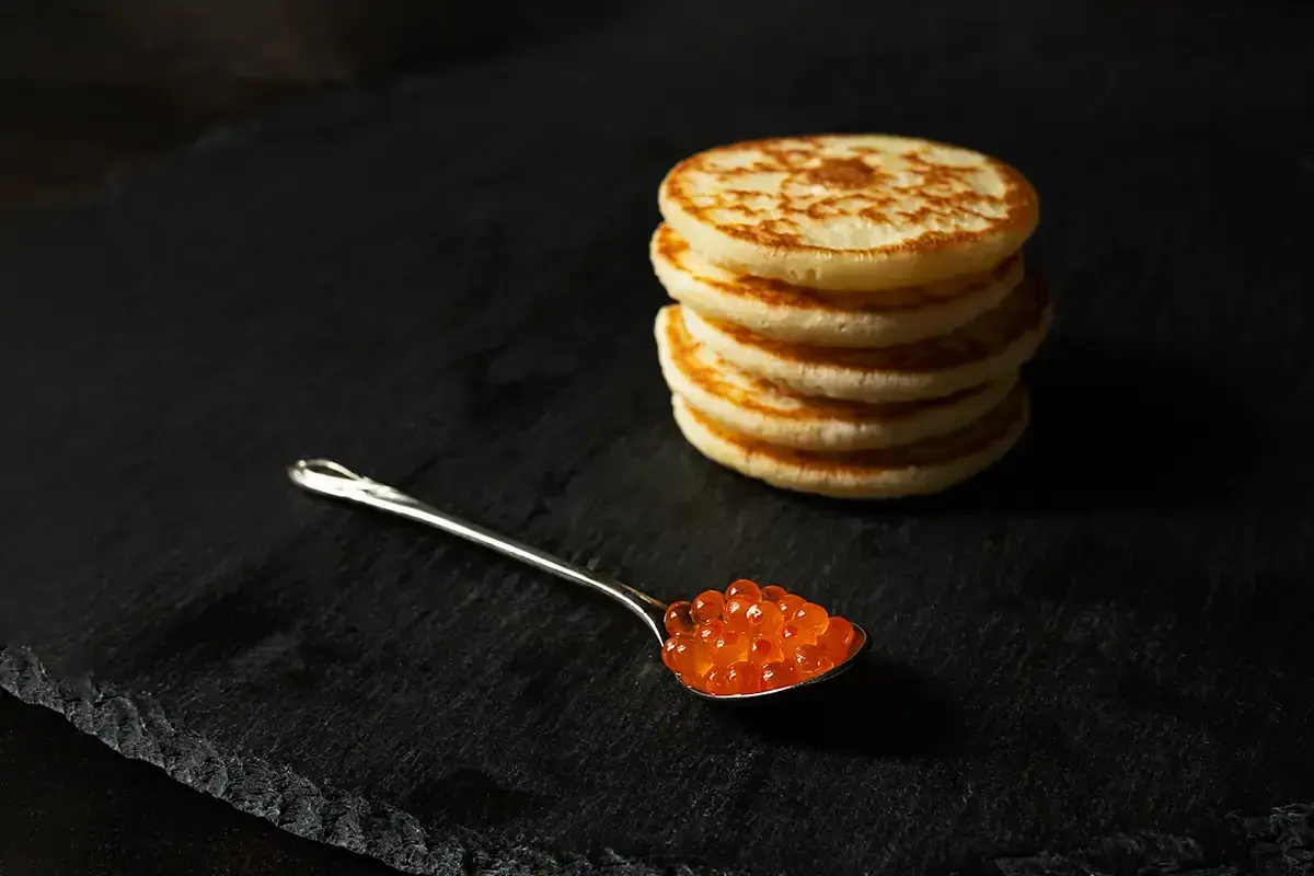 Une cuillère de caviar rouge sur une assiette noire, à côté d'une pile de blinis dorés. Parfait pour une recette blinis gourmande.