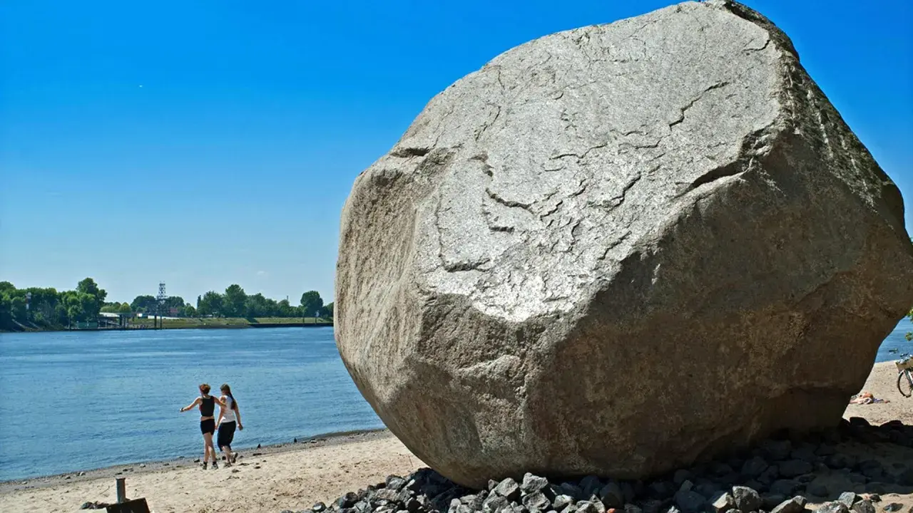Der alte Schwede, ein riesiger Findling, thront am Ufer. Zwei Frauen spazieren am Wasser entlang, im Hintergrund Bäume und ein blauer Himmel.